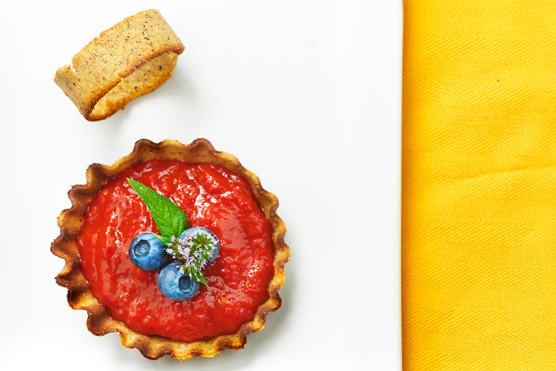 Questo dessert è nato dall'esigenza di fare delle prove per il dolce da presentare al Joia Academy per l'esame. Fin dal principio avevo ben focalizzato quale dessert preparare, che sarebbe stato interamente crudista è suddiviso in tre parti. Una simil panna cotta ai lamponi su brownies di cacao e noci, una crema pasticciera crudista allo zafferano con caviale d'anguria, pistacchi e liquirizia e una sfoglia croccante di lino e banana con un gelato crudista alla pesca. Il crudismo è per me una tranquillità. Mi sento abbastanza ferrata e mi muovo con tranquillità. Conosco gli ingredienti e le tecniche. So come utilizzare la frutta e i semi così come ingredienti più particolari come l'alba irish moss o la xantana. Giocavo quindi su un terreno sicuro, una mia certezza. Se nel primo e secondo dessert ero assolutamente convinta però, del terzo non ero fermamente convinta. Ogni giorno che passava, e ad ogni lezione, quella certezza di fare tutto crudo vacillava. Cosa avrei imparato se restavo ferma sulle mie conoscenze? Uscire dalla mia zona di comfort non mi faceva paura ma mi aveva lasciato il vuoto nel terzo dessert che componeva il piatto. Se non avessi fatto una cialda con il gelato, cosa avrei potuto fare? Forse era più probabile fare qualcosa di più elaborato ma mantenendo l'idea iniziale. Un giorno lessi per caso una frase tratta dal libro di "Alice nel paese delle meraviglie", uno dei miei libri preferiti ed il primo che lessi da sola da bambina: . Lessi più volte questa frase è tutto mi fu chiaro. Come una illuminazione, come se nella testa avesse iniziato a suonare un campanello. "Tutto dipende da dove vuoi andare". Certo, se io fossi Alice, vorrei andare incontro al Bianconiglio. Ognuno di noi ha il proprio Bianconiglio. Il mio dessert non era " questa sono io" ma "dove sono e dove sto andando". Così era più appropriato! E dove starei andando? Verso un mondo fatto di mestoli e stoviglie, di dessert peccaminosi e opere d'arte nel piatto. Caspita, sto studiando dai più grandi in assoluto e gli presento c'è cosa? Il vortice nel mio cervello aveva iniziato a riprendere velocità. Avrei comunque fatto una cialda ma doveva essere più particolare. Il gelato crudista è così diventato il gelato alla mela che avevo visto al Vegetarian Change. Lo chef che lo aveva preparato, in versione salata alle ortiche, al termine della sua performance si era gentilmente fermato a parlare con me. Ne avevo approfittato per farmi raccontare il piatto che mi aveva particolarmente colpito. Io sono come San Tommaso ed avevo provato a rifarlo. Mi aveva precisato che, grazie alla pectina contenuta nella mela, una volta montato il gelato non si scioglie. Ed era assolutamente vero. Sarebbe stato il mio ripieno. Una mela cotta alla cannella, trasformata in gelato. Era comunque un gelato, come lo avevo immaginato all'inizio, ed era una mela cotta, in assoluto il mio dessert preferito. Io amo i dolci semplici e semplice doveva comunque rimanere anche il piatto che avrei presentato. Il guscio che l'avrebbe contenuto doveva essere friabile, leggero e lo volevo senza glutine e lievito, così da poterlo mangiare anche io. In questi mesi ho lavorato molto con la farina di ceci, studiandone le caratteristiche e provando a realizzare diverse preparazioni. L'ho unita alla farina di grano saraceno che, oltre a darmi quel tocco di rusticità della Taverna, mi permetteva di dare robustezza e consistenza alla preparazione. Avevo imparato che, utilizzando acqua bollente, si creava una maglia elastica che permetteva di lavorare la farina. Ho sfruttato quindi questa conoscenza, oltre ad aggiungere lo sciroppo d'agave, per ottenere una frolla friabile e lavorabile. Le prima prove mi avevano soddisfatto in termini di impasti che avevo modellato a corolla. Sembravano le gonne di tulle delle ballerine, vaporose e allegre e, nonostante mi piacessero e mi rappresentassero molto, sapevo che non erano giuste per la mia presentazione. Pensando e ripensando alla fine ho scelto una forma cilindrica che più si addiceva al piatto nel suo complesso. Il piatto risultava equilibrato. Un percorso, come il mio di studio, che mi portava dal dessert crudista super collaudato a una tartelletta farcita con gelato di mela, rinfrescante, e pesca caramellata alla limoncina. Nonostante lo stravolgimento del piatto, avevo però mantenuto la costante della semplicità e della "rusticità" che contraddistingue la Taverna. Per dare una nota caratteristica al piatto ho concluso decorando con caviale di anguria. Il caviale ho imparato a farlo studiando la cucina molecolare. Una cucina che mi diverte molto e che mi piace sperimentare. Non sono sicura che vorrei mangiare un piatto, o un'intera cena, di questo tipo di cucina ma aggiungere dei dettagli ai piatti lo trovo gradevole. La cucina molecolare mi costringe ad analizzare più attentamente la chimica degli alimenti e degli elementi utilizzati e lo trovo molto interessante. Al termine delle mie prove avevo diverse tartellette, di varie forme e misure, che non sapevo come usarle. Con l'unico peperone che mi era rimasto in frigo ho preparato una salsa dolce. Ho aromatizzato con le mie amate spezie e cotto tutto con sciroppo d'agave fino ad ottenere una consistenza simile alla crema pasticciera. Un'altra cosa interessante che ho imparato al Joia è di osare e non relegare gli ortaggi alle preparazioni salate e la frutta a quelle dolci ma di utilizzarle a 360 gradi. A Piccolo Chef, che adora i peperoni, è piaciuta molto e sono finite in un lampo. Frolla ai ceci con crema pasticciera ai peperoni